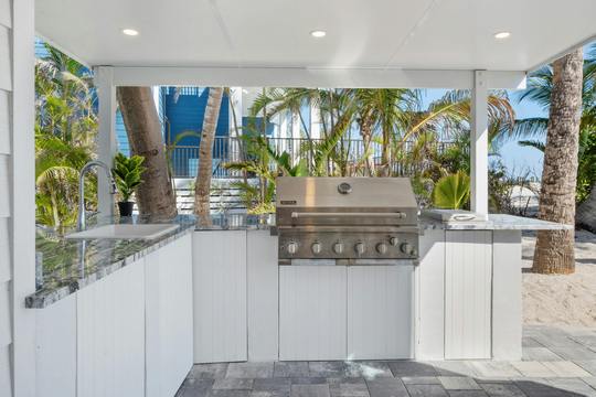Modern outdoor kitchen with a grill and countertop, featuring palm trees and a pool in the background.